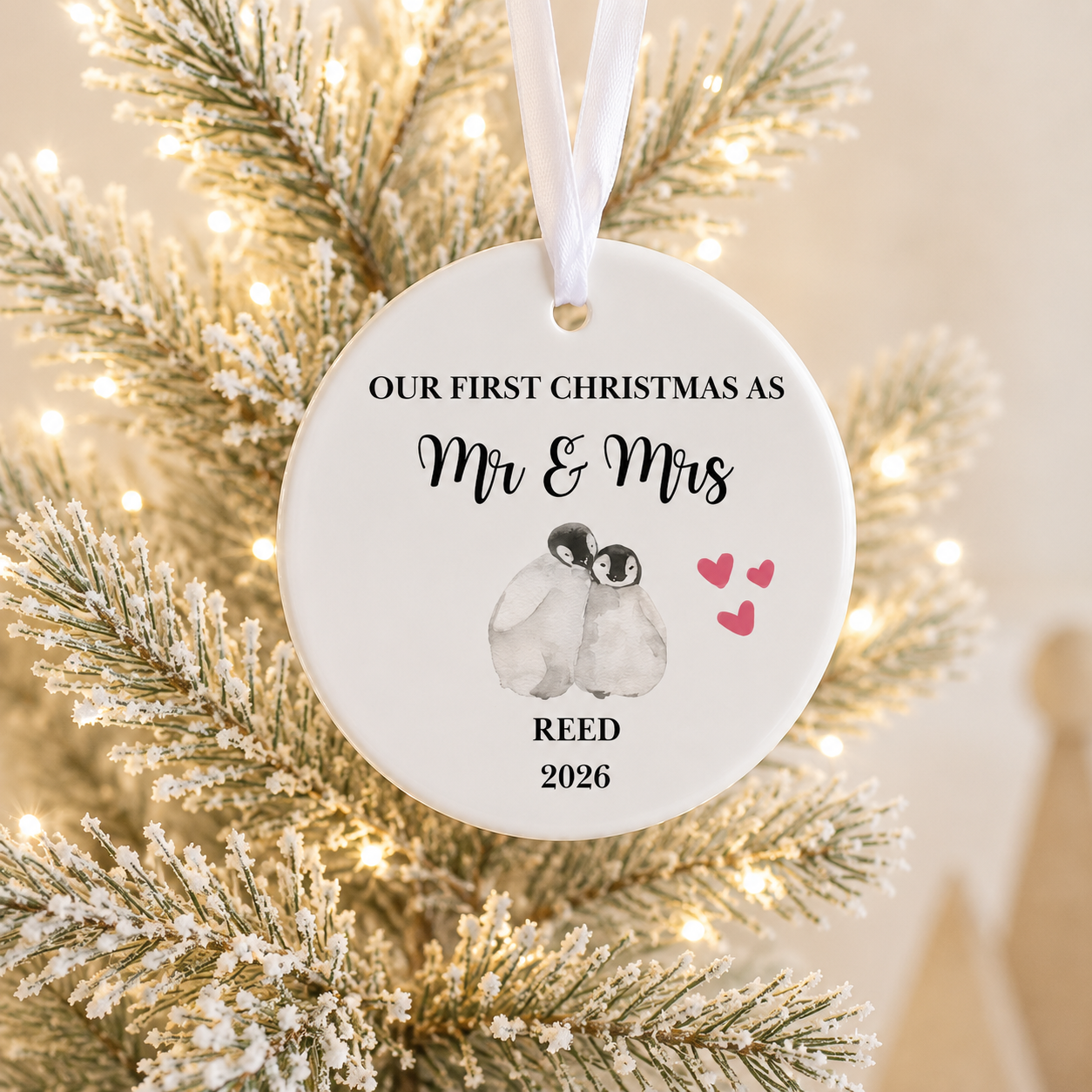 A round white personalized Christmas ornament hanging from a white ribbon, printed with the text 'OUR FIRST CHRISTMAS AS Mr & Mrs REED 2026' and an illustration of two penguins with pink hearts, shown against frosted tree branches with warm string lights.