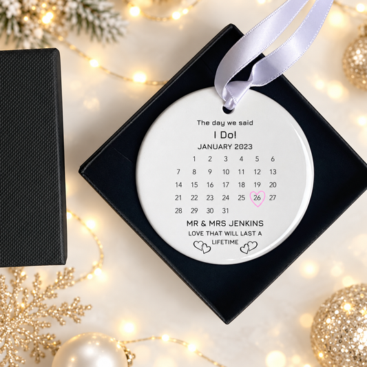 A personalized round hanging ornament displayed in a black square gift box, with a white ribbon loop at the top. The ornament is printed with a calendar for January 2023 and the text 'The day we said I Do!' and 'MR & MRS JENKINS LOVE THAT WILL LAST A LIFETIME' with a pink highlighted date.