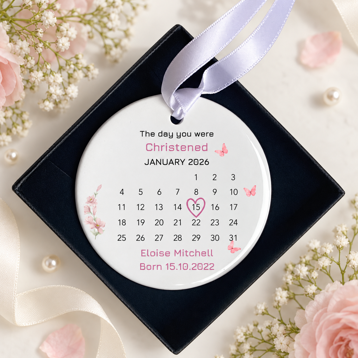A personalized round Christened birth calendar ornament displayed inside a black square gift box, decorated with pink roses, baby's breath, and a white satin ribbon.