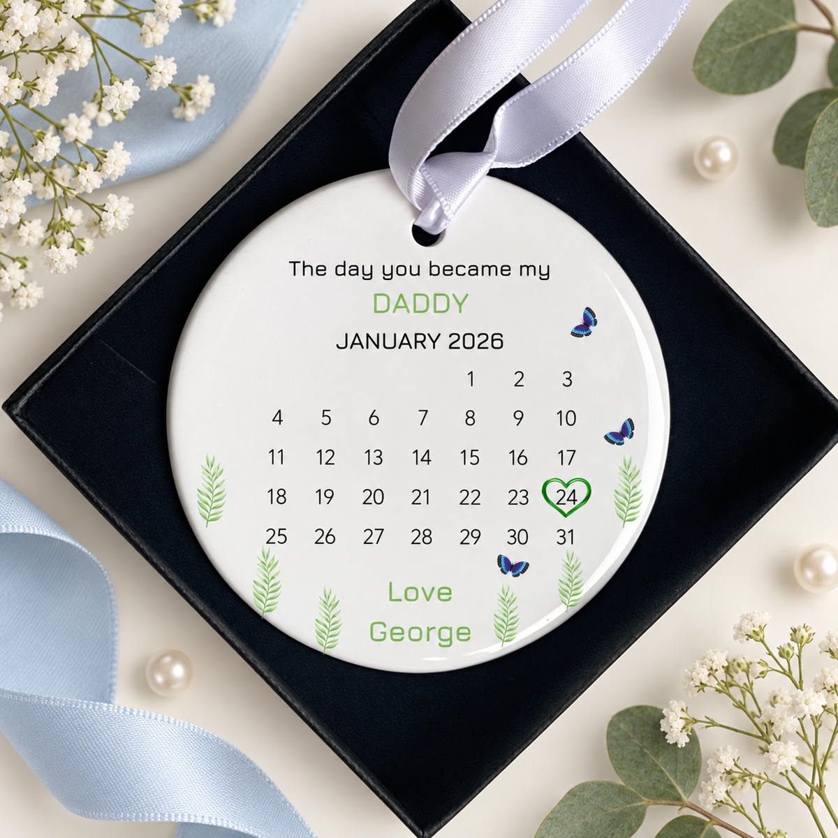 A personalized round white Christmas ornament displayed inside a black square gift box, decorated with a white satin ribbon. The ornament features a printed January 2026 calendar with the text 'The day you became my DADDY', the number 24 highlighted in green, and 'Love George' at the bottom.