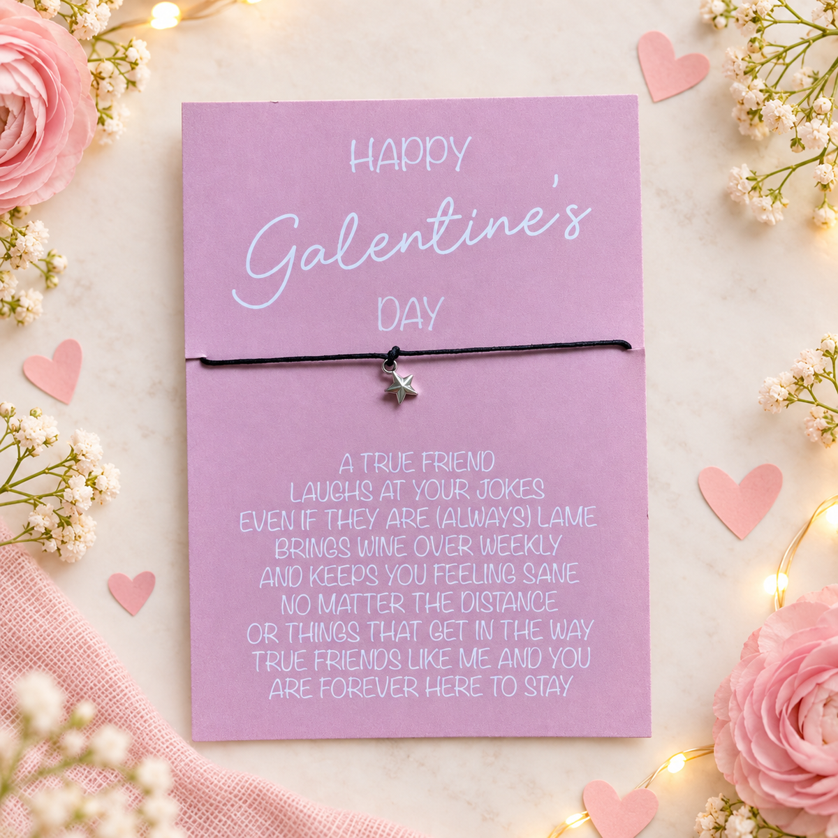 A pink Valentine's Day greeting card with white text and a black cord bracelet featuring a small silver star charm, styled with pink roses, baby's breath, and fairy lights.