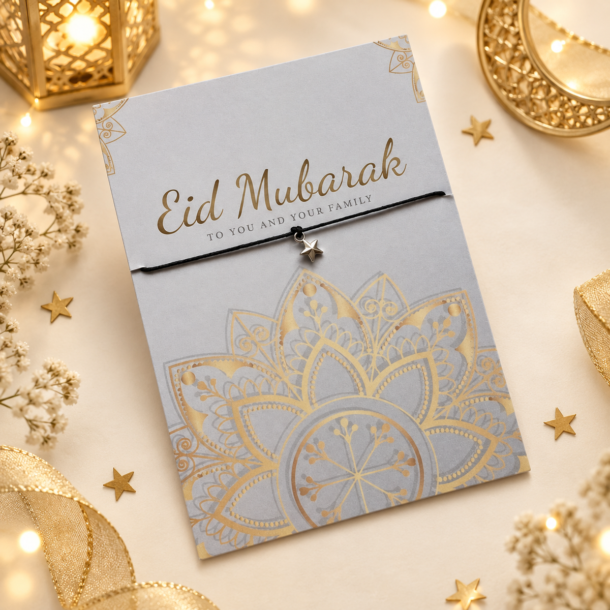 An Eid Mubarak wish bracelet card with a gold foil mandala design and a small star pendant, styled on a warm cream surface with decorative gold lanterns, stars, and dried flowers.