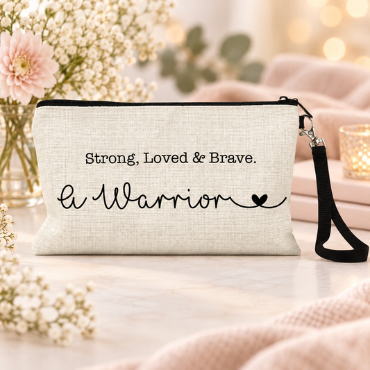 A beige woven clutch or wristlet bag with a black zipper, black wrist strap, and black printed text reading 'Strong, Loved & Brave. A Warrior' on a soft pink and white styled tabletop scene.