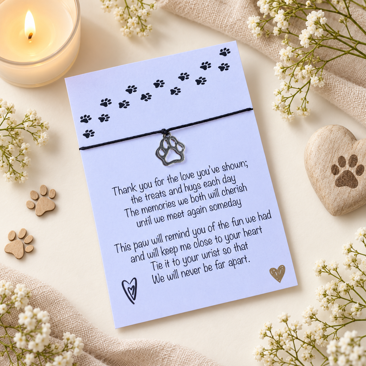 A pet memorial card with a black cord and paw print charm, styled in a flat lay with a lit candle, white flowers, wooden paw prints, burlap fabric, and a heart-shaped stone.