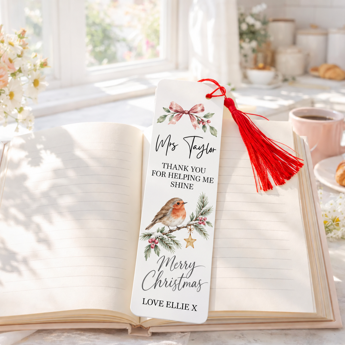 A personalized Christmas bookmark with a red tassel, featuring a robin bird, green holly leaves, and a pink bow, displayed on an open book in a bright, sunlit room.