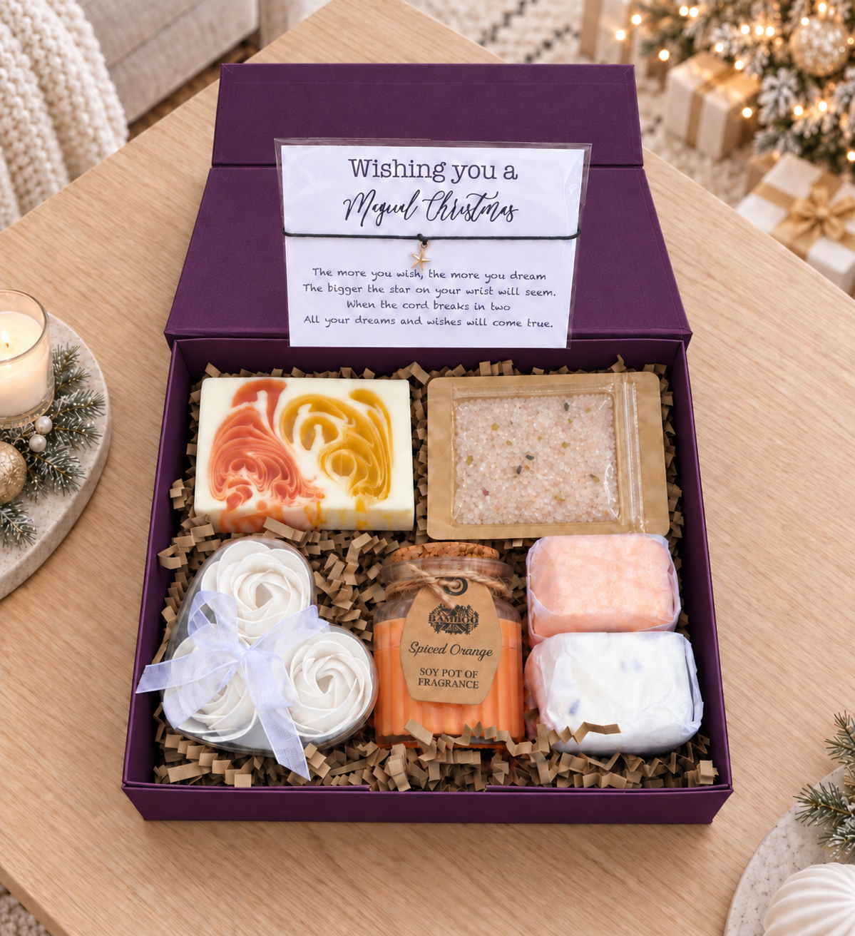 A holiday gift box set on a wooden table, featuring a purple box with a 'Wishing you a Magical Christmas' card, containing artisanal soaps, a bath salt packet, and a candle.