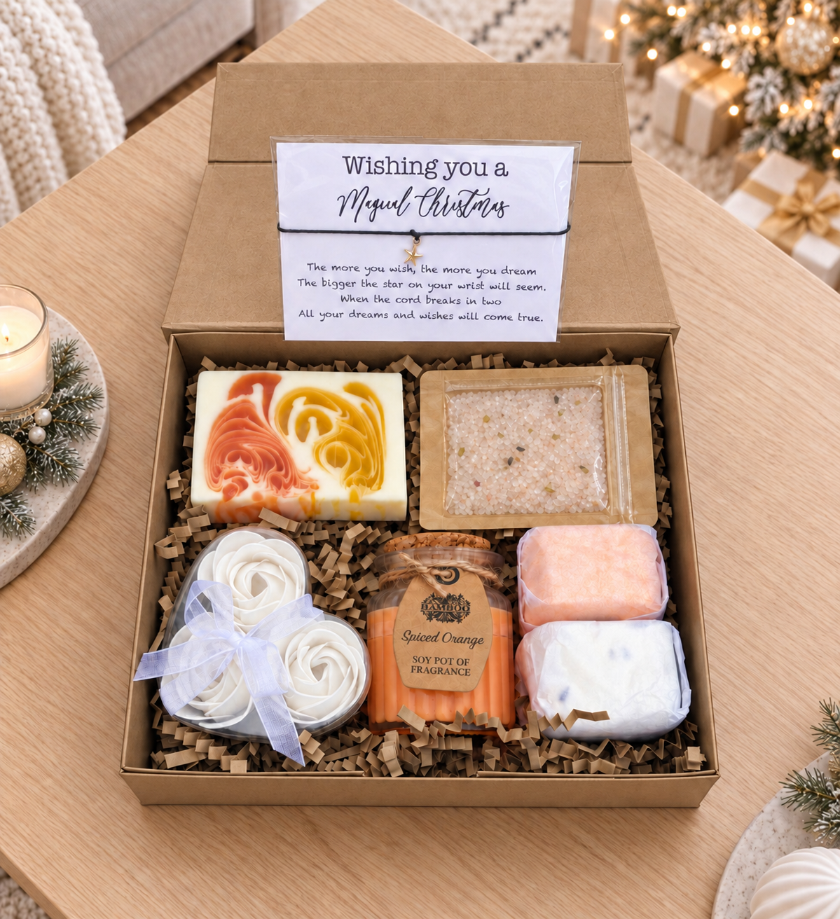 A gift box of handmade soaps and bath items displayed on a wooden table, with a candle and a decorated Christmas tree in the background. The open kraft box contains a marbled soap, a bath salt packet, a rolled white soap tied with a ribbon, and two shower steamers, with a card reading 'Wishing you a Magical Christmas' inside.