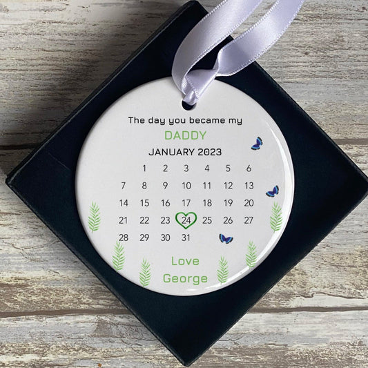 Personalised ‘The Day You Became My Daddy’ ceramic ornament featuring a calendar for January 2023, with the date 24th marked by a heart. Decorated with green leaves and blue butterflies, making it a unique and meaningful gift for a new father.