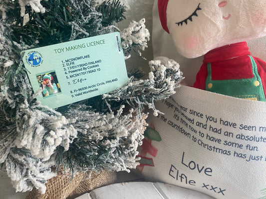 A close-up of a novelty "Toy Making Licence" card nestled in the snowy branches of a small artificial Christmas tree. The green card features playful details including the name "McSnowflake," the elf name "Elfie," a birthdate listed as "17/01/190AD FINLAND," and the phrase "Powered by Cookies." The license includes a small photo of an elf and a signature, giving it a whimsical, official look. Perfect for Christmas elf arrival props or festive play.