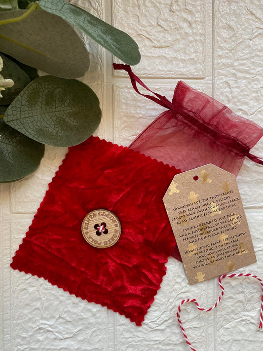 A red velvet pouch with a sewn button resembling Santa's button, placed next to a small organza bag and a gift tag with a Christmas message.