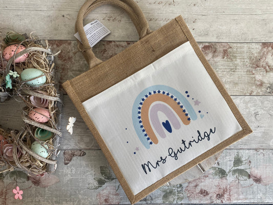 Custom jute tote bag with a pastel rainbow design in peach, lavender, and light blue tones, surrounded by navy stars. The name "Mrs Gutridge" is printed underneath in a flowing handwritten-style font. Decorative Easter wreath visible on the side.