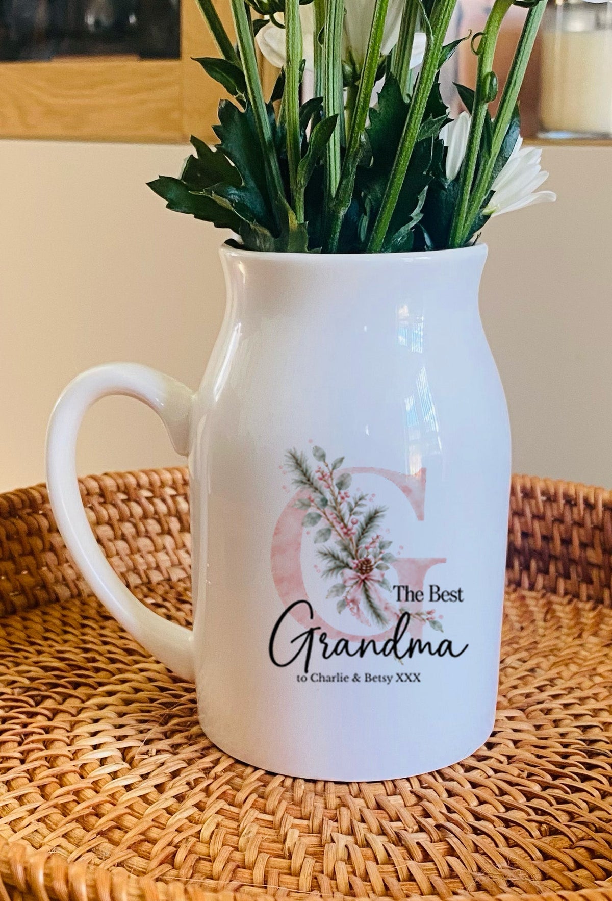 A white ceramic pitcher vase with a floral design and the words 'The Best Grandma' on the front, filled with white flowers, sitting on a round woven rattan tray.