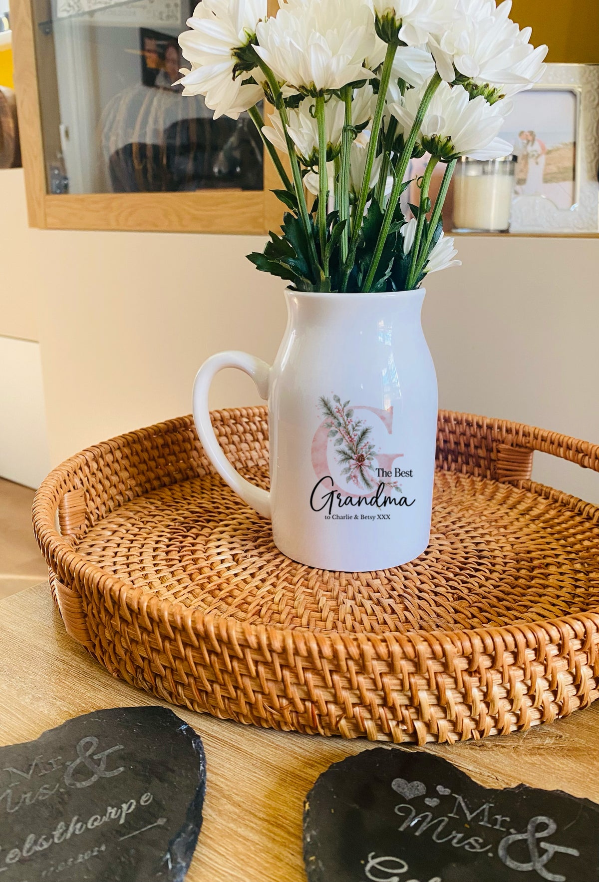 A white ceramic pitcher vase with a floral design and the words 'The Best Grandma' on the front, filled with white flowers, sitting on a round woven rattan tray.