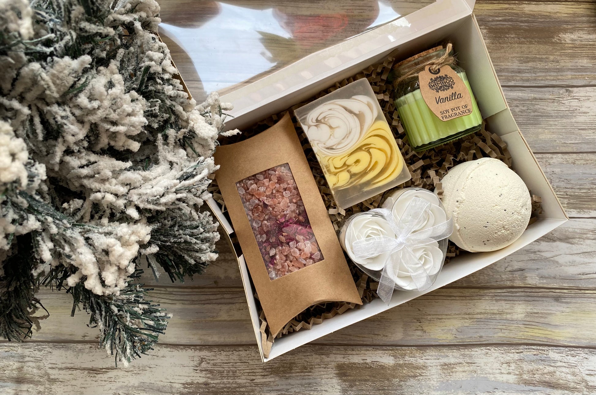 Gift box with bath products on a wooden surface next to a decorative tree.