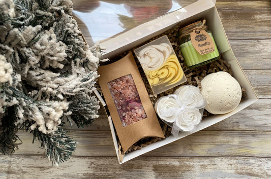 Gift box with bath products on a wooden surface next to a decorative tree.