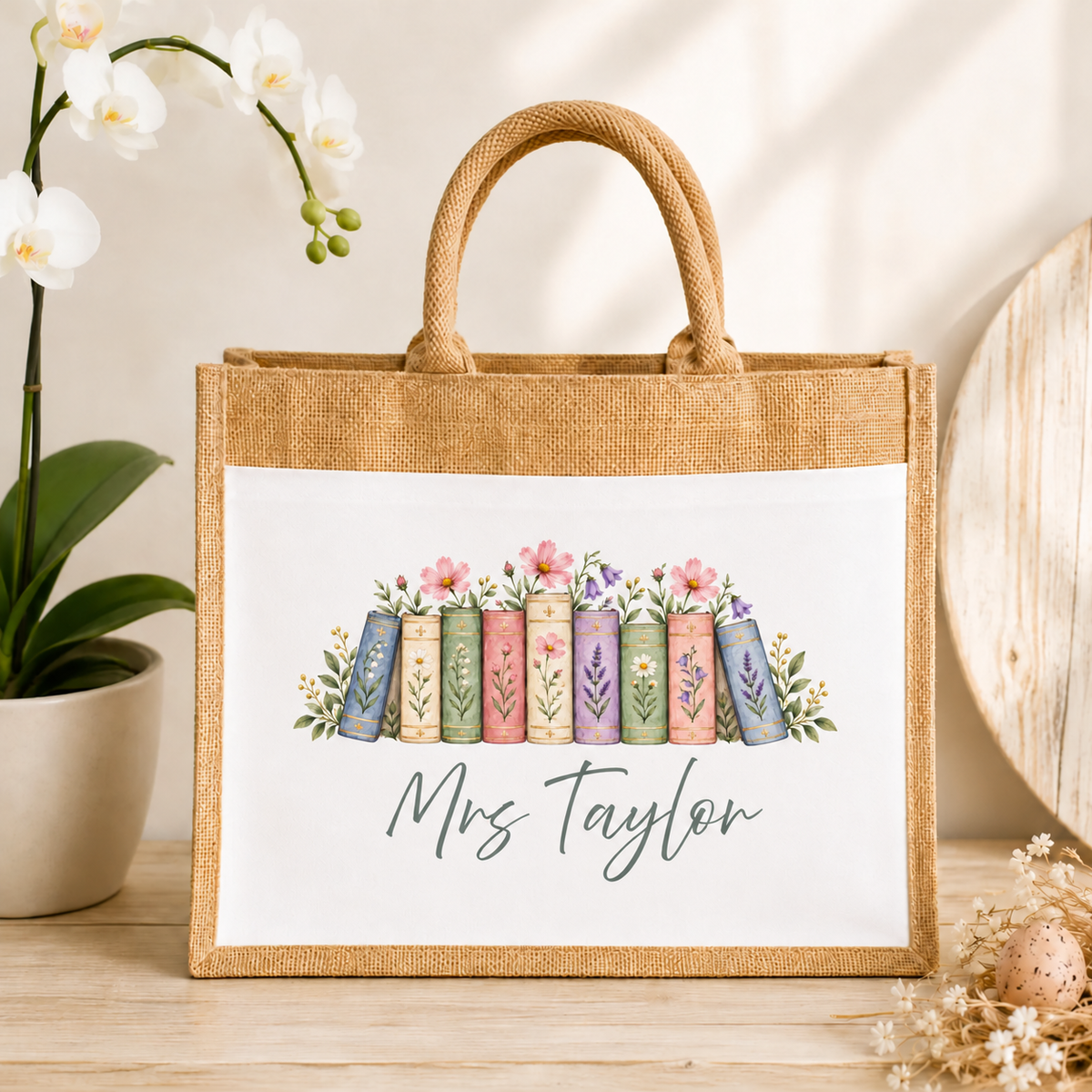 A personalized jute tote bag with rope handles, featuring a white front panel printed with a row of colorful vintage books topped with flowers and the name 'Mrs. Taylor' in script.