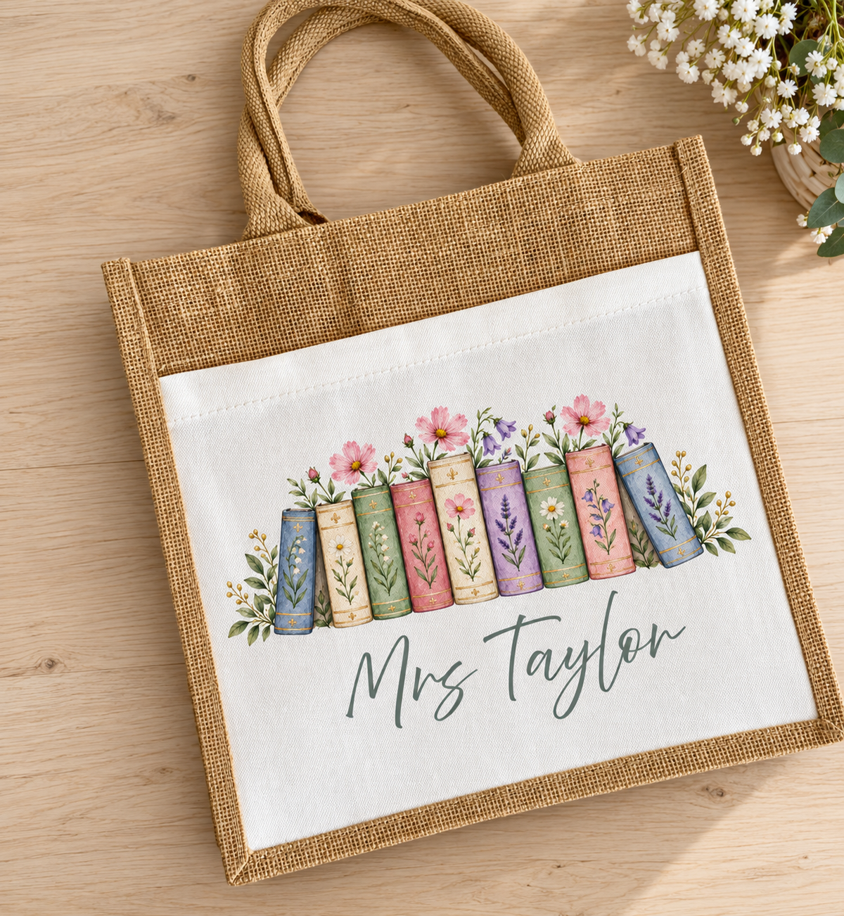 A personalized jute tote bag with a white printed panel showing a row of colorful book spines, flowers, leaves, and the name 'Mrs Taylor' in script, photographed on a light wood surface with small white flowers in the corner.