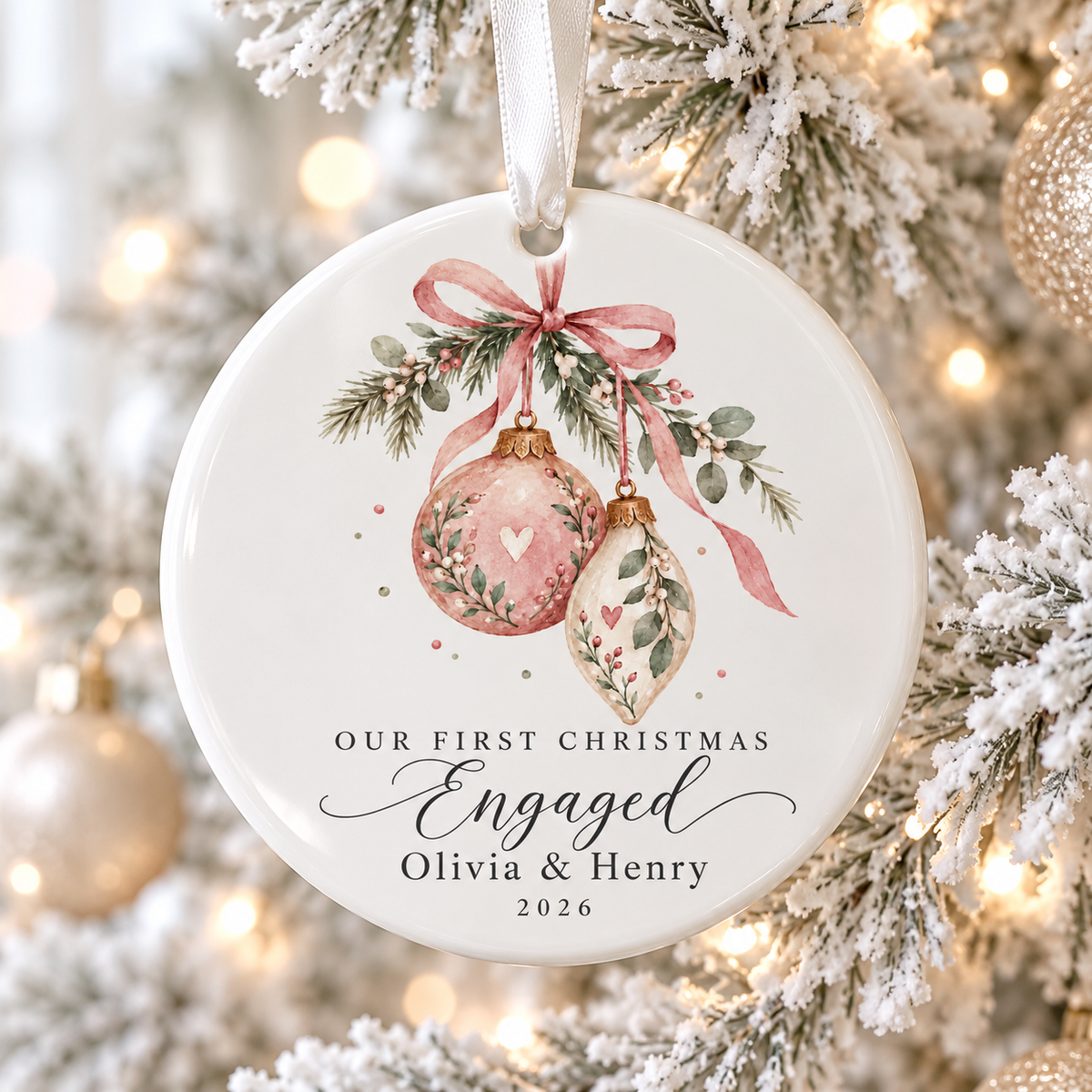 A round white ceramic Christmas ornament hanging from a white ribbon on a decorated tree. The ornament features a watercolor design of two pink and cream floral ornaments under a pink ribbon, with the text 'OUR FIRST CHRISTMAS Engaged Olivia & Henry 2026'.