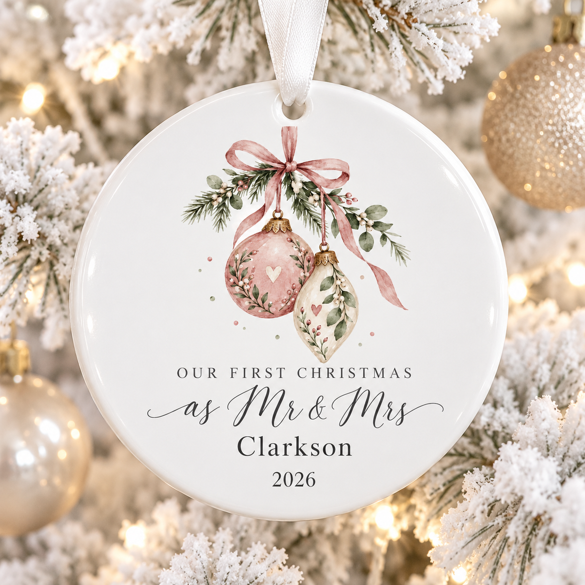A round white ceramic Christmas ornament hanging from a white ribbon, printed with the phrase 'Our First Christmas as Mr & Mrs Clarkson 2026' beneath two pink and gold ornaments with green foliage and a pink bow. The ornament is photographed against a festive blurred Christmas tree background with warm lights and gold ornaments.