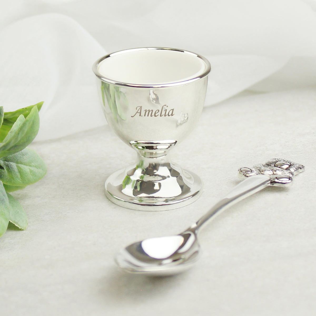 Silver egg cup and spoon with 'Amelia' engraving on a white background
