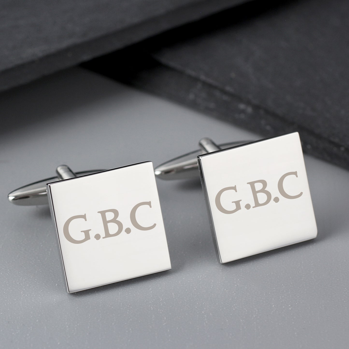 Square cufflinks with 'G.B.C.' engraving on a gray surface