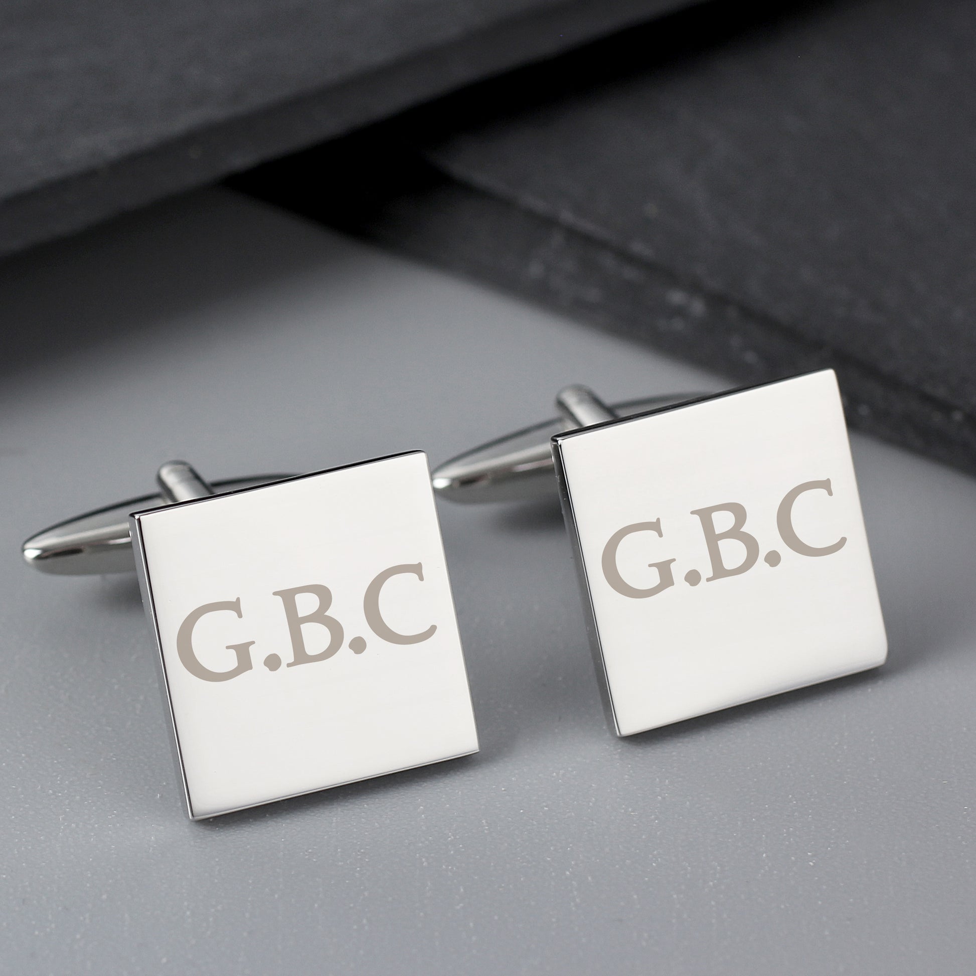 Square cufflinks with 'G.B.C.' engraving on a gray surface