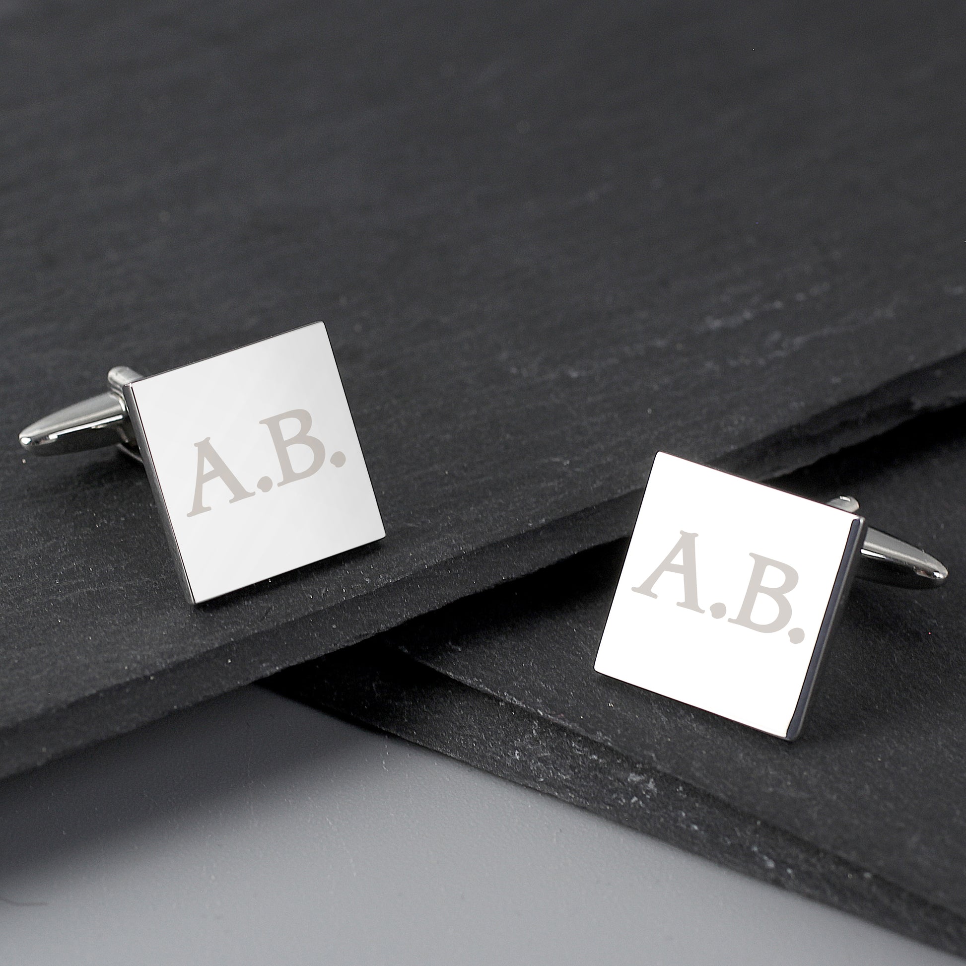 Square cufflinks with 'A.B.' engraving on a dark surface