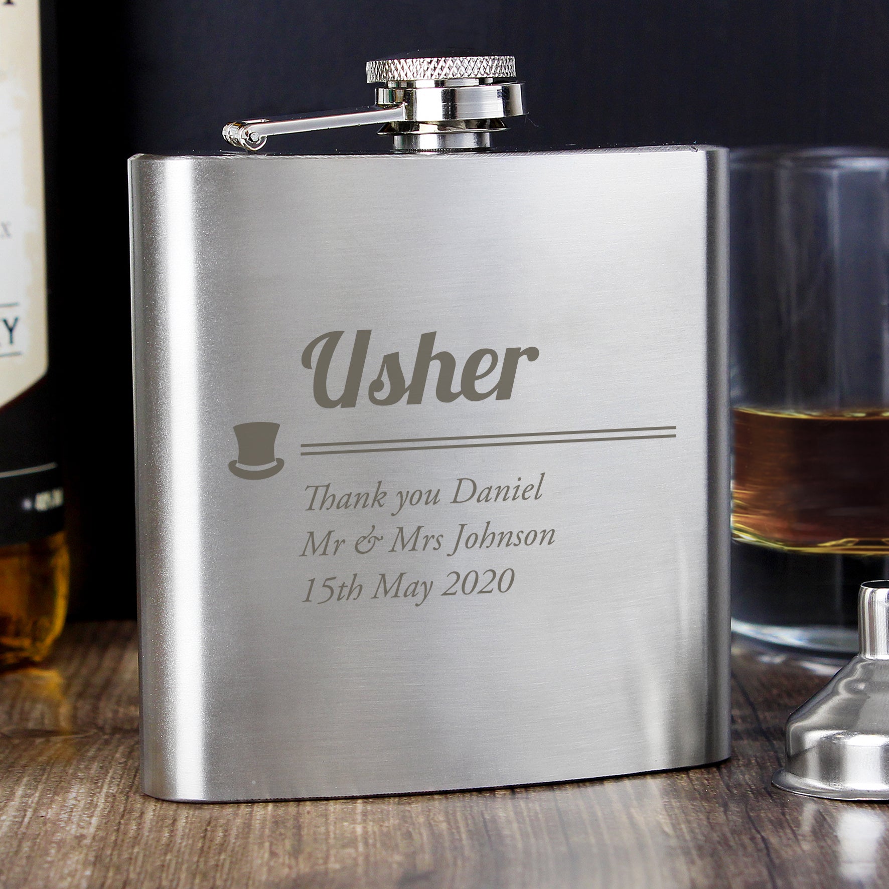 Silver flask engraved with 'Usher' and personalised text, placed on a wooden surface with a glass of whiskey in the background.