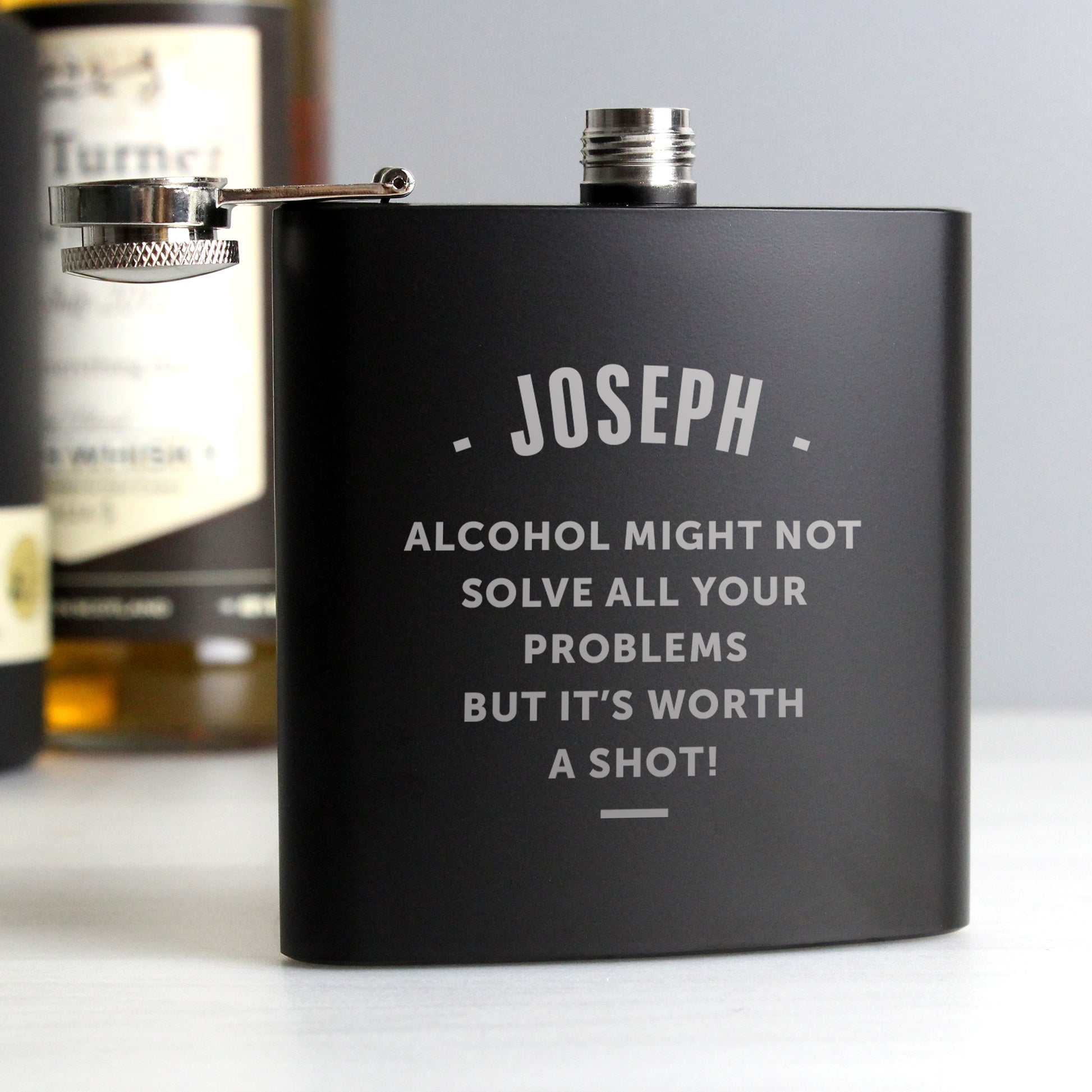Personalised Black flask with 'JOSEPH' and humorous text, placed on a surface with bottles in the background.