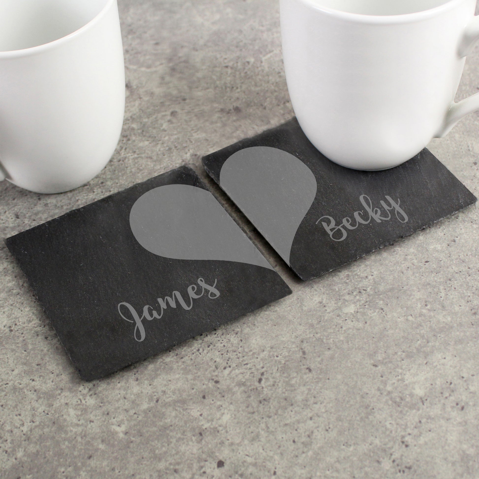 Two slate coasters with grey heart designs and names 'James' and 'Becky' on a concrete surface.