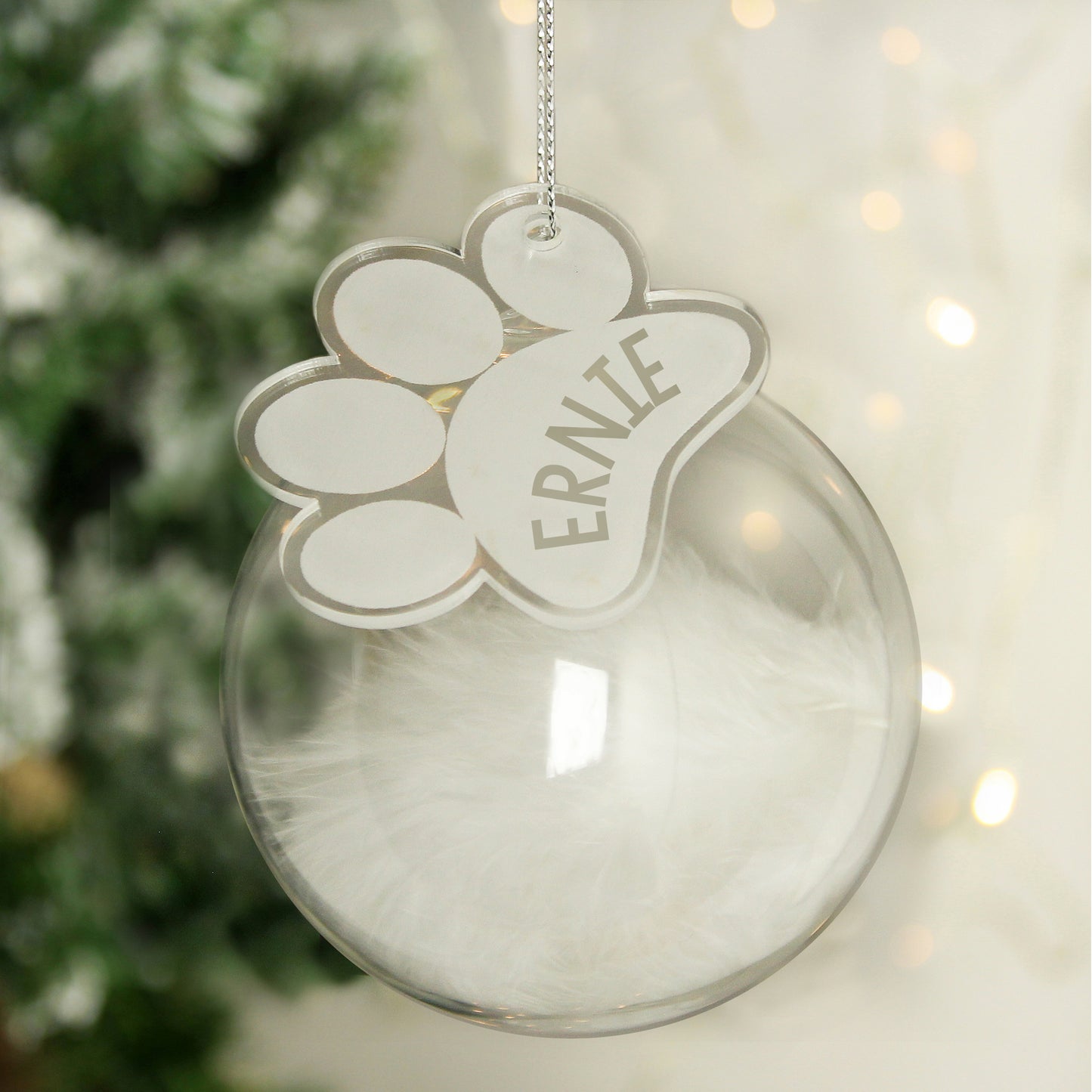 Clear Christmas ornament with a paw print design and 'ERNIE' text, hanging against a blurred festive background.