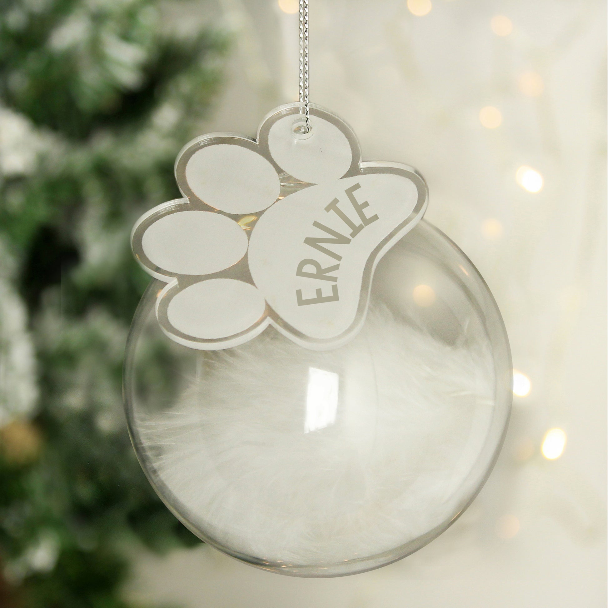 Clear Christmas ornament with a paw print design and 'ERNIE' text, hanging against a blurred festive background.