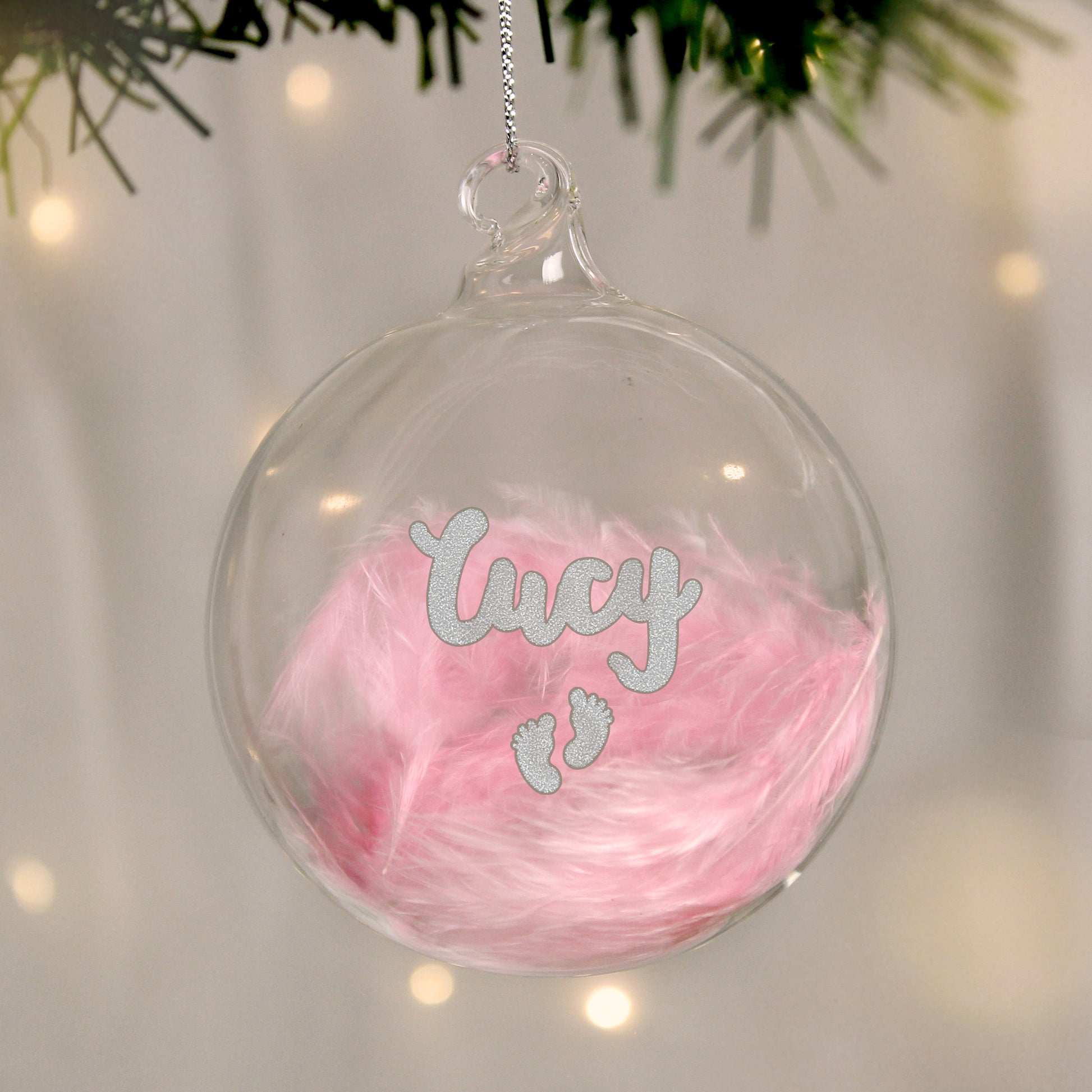 Clear glass ornament with pink feathered interior and 'Lucy' and footprints in silver, hanging against a blurred festive background.