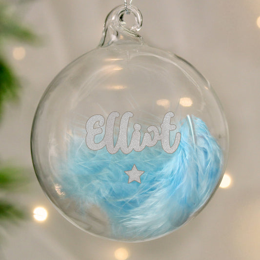 Clear glass ornament with blue feathers and 'Elliot' text, hanging against a blurred background.