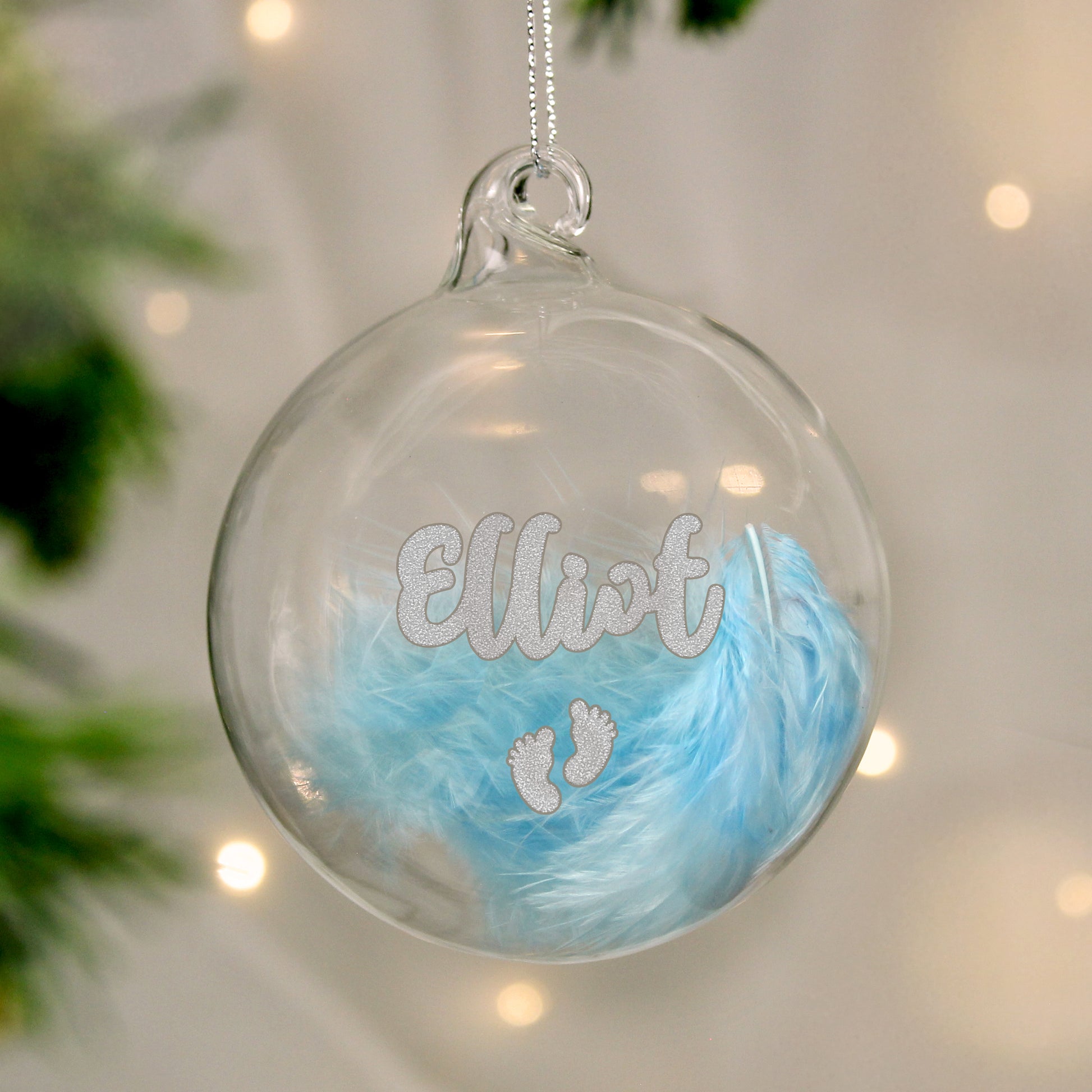 Clear glass ornament with blue feathers and 'Elliot' text, hanging against a blurred background.
