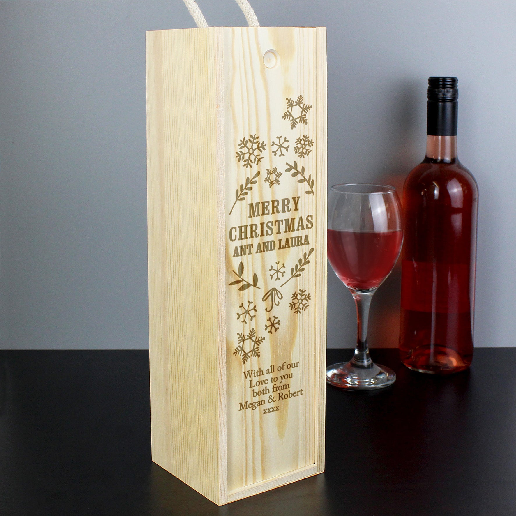Wooden wine box with engraved text, next to a bottle of red wine and a glass on a dark surface.