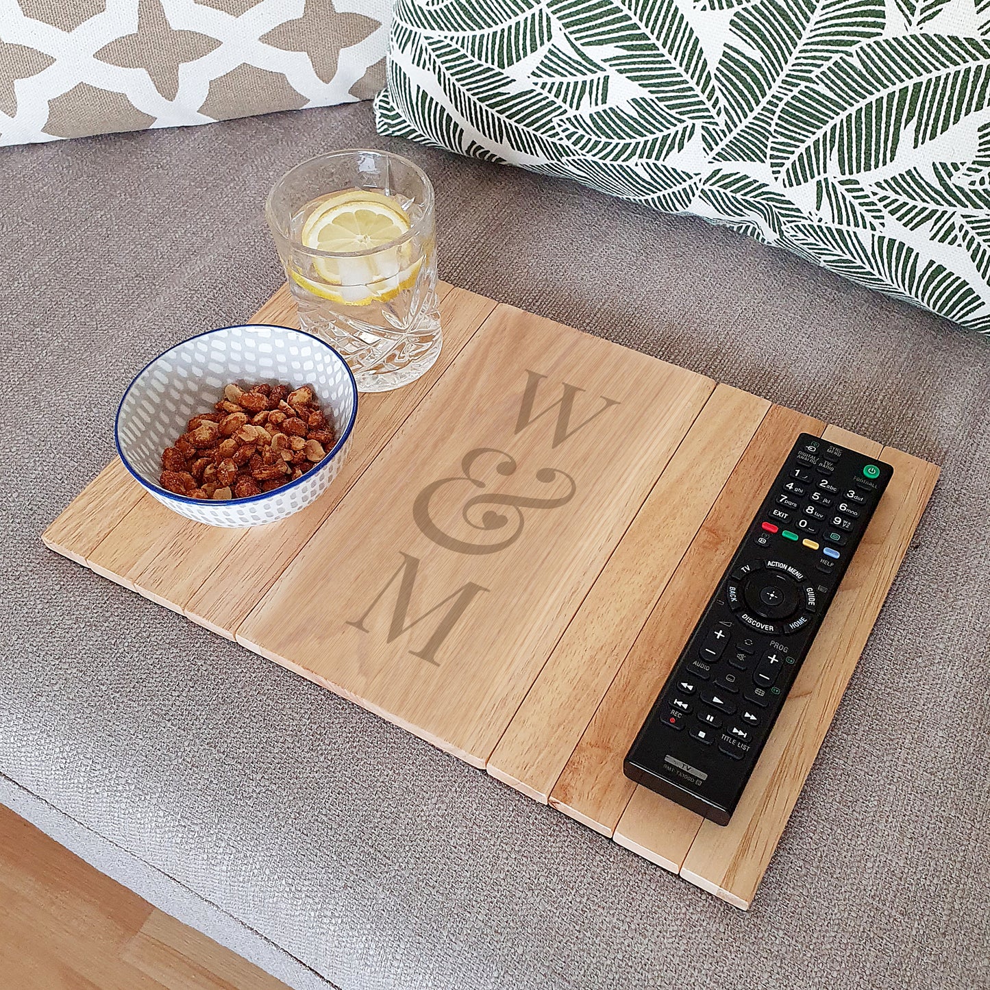 Wooden tray with 'W & M' engraving, bowl of snacks, glass of water, and remote control on a couch.