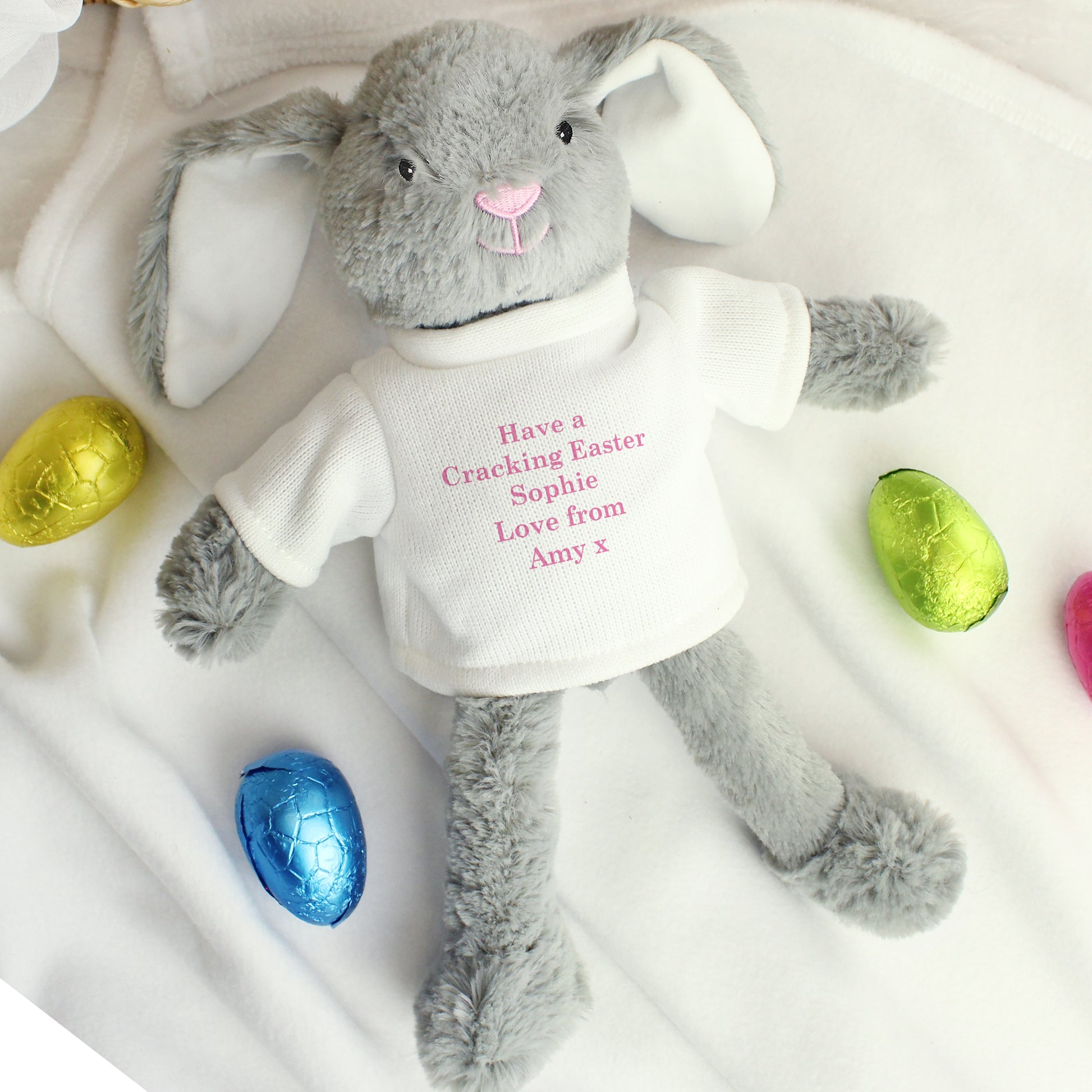 Grey plush bunny with a white shirt displaying text, surrounded by colourful Easter eggs on a white background