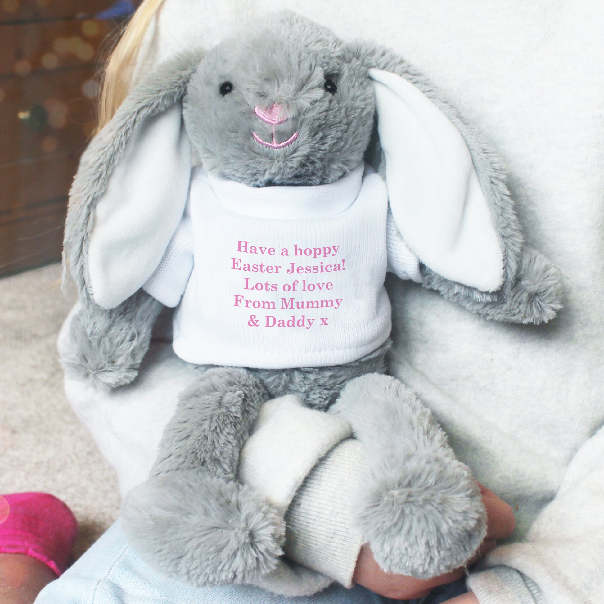 Grey plush bunny with a white shirt displaying personalised text, held by a person.