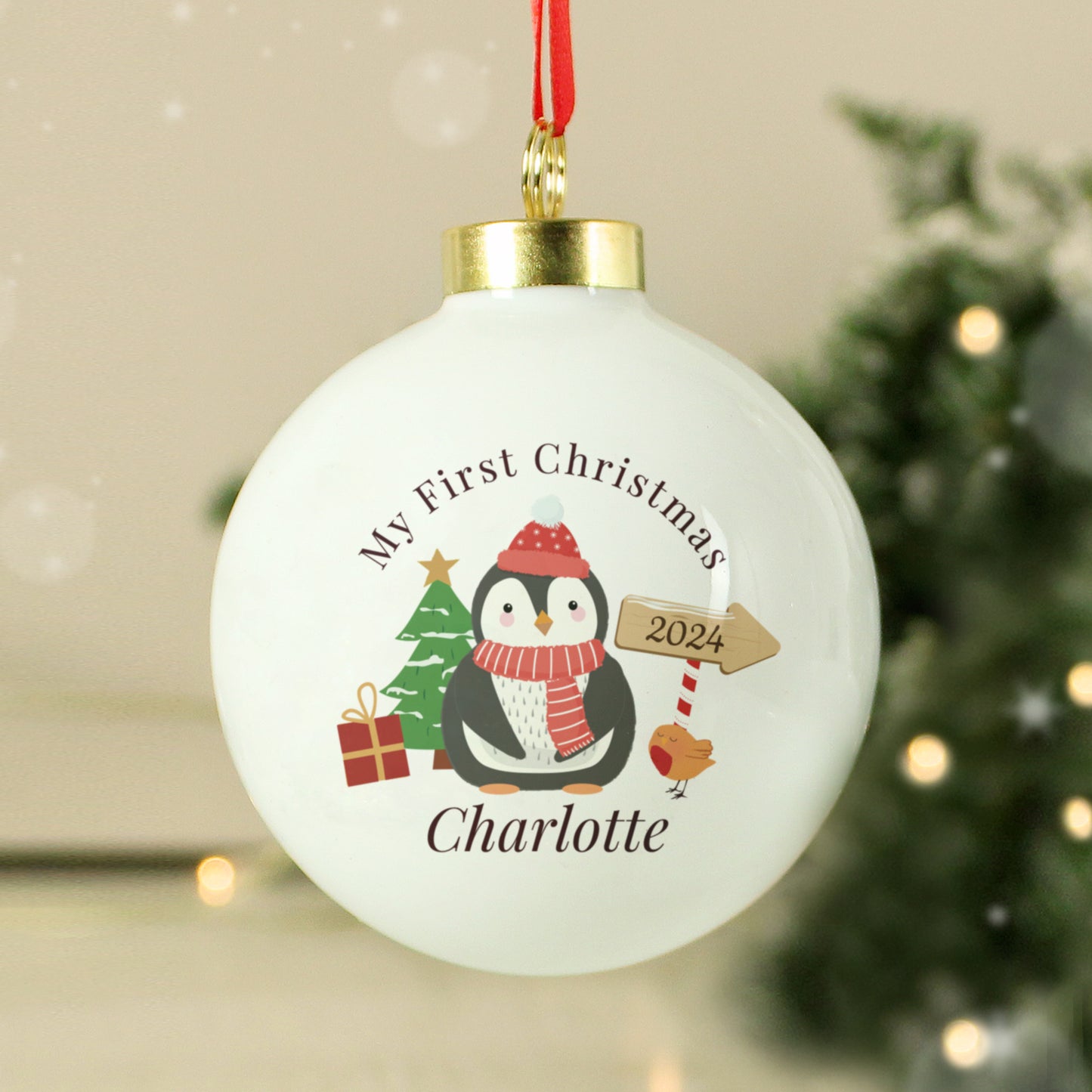 Christmas ornament with penguin design and text 'My First Christmas 2024 Charlotte' on a blurred festive background.