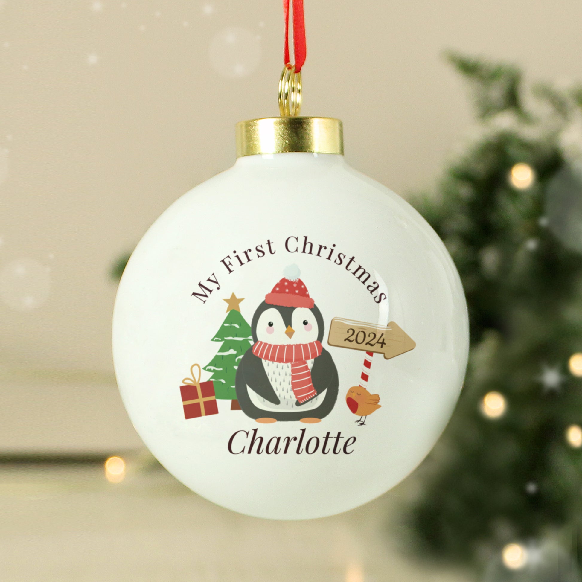 Christmas ornament with penguin design and text 'My First Christmas 2024 Charlotte' on a blurred festive background.