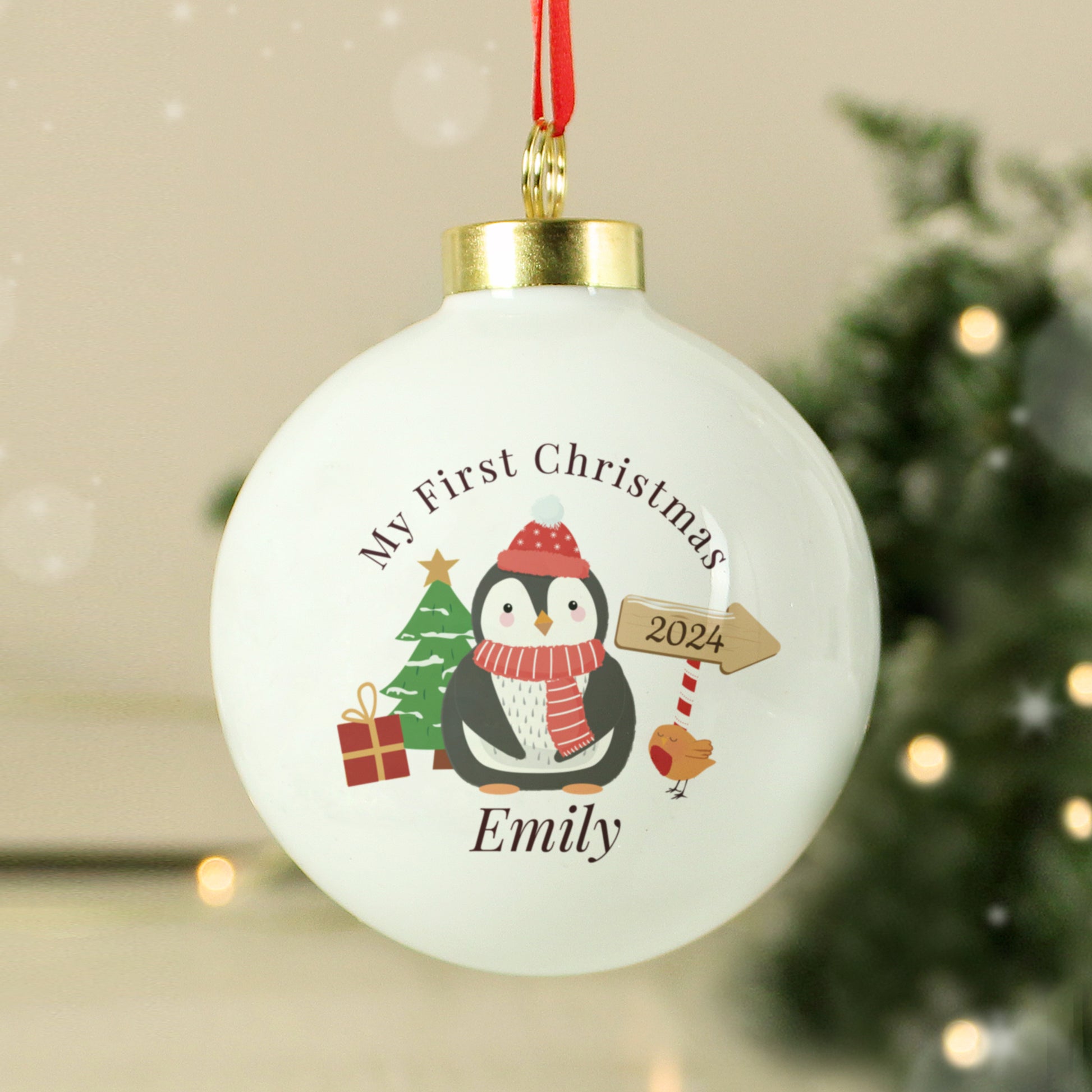 Christmas ornament with penguin design and text 'My First Christmas 2024' on a blurred festive background.