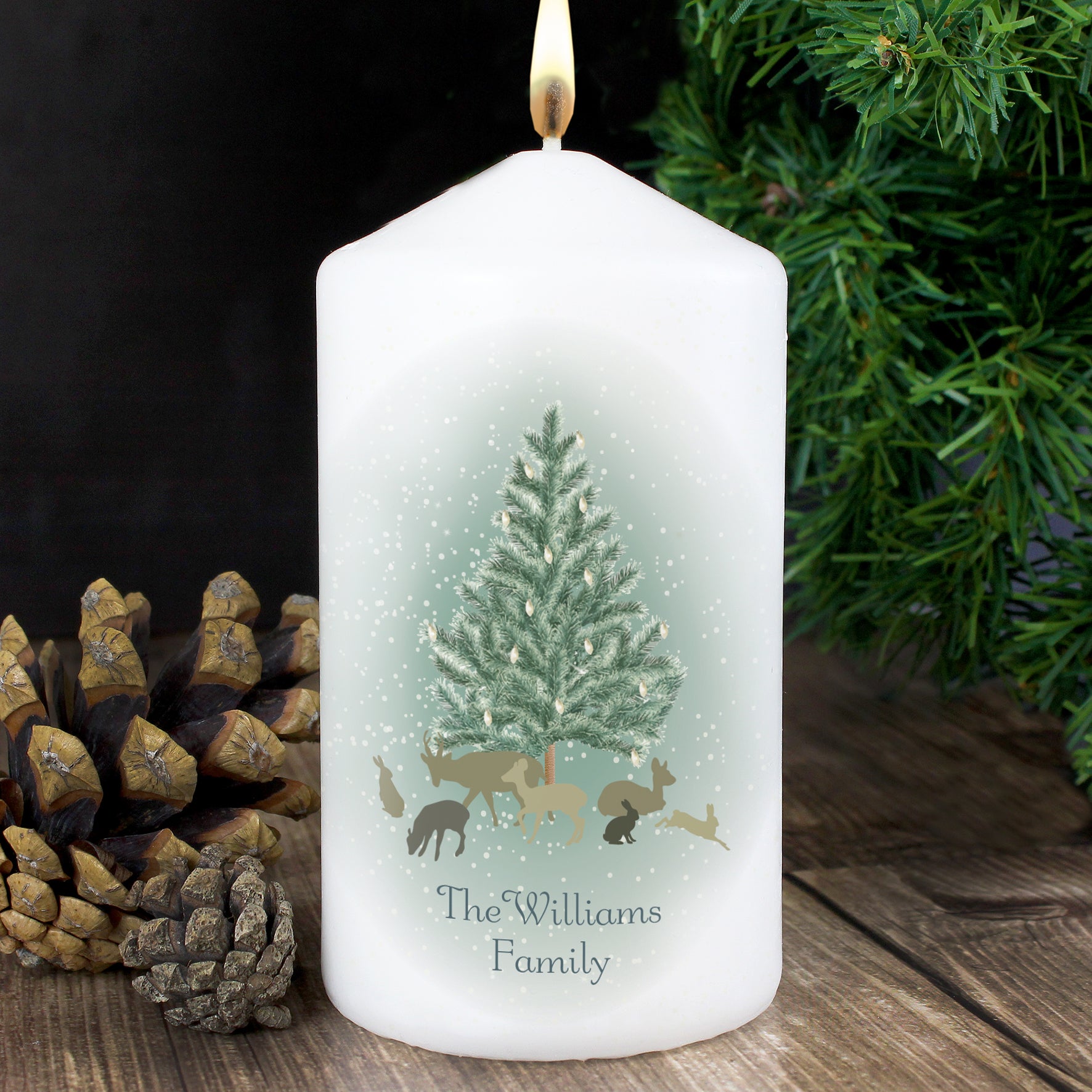 White candle with a Christmas design featuring a tree and animals, placed on a wooden surface with pinecones and greenery.