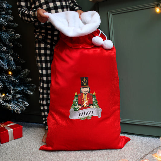 Red Christmas sack with a nutcracker design and 'Ethan' text, held by a person in checkered pajama pants next to a decorated tree.