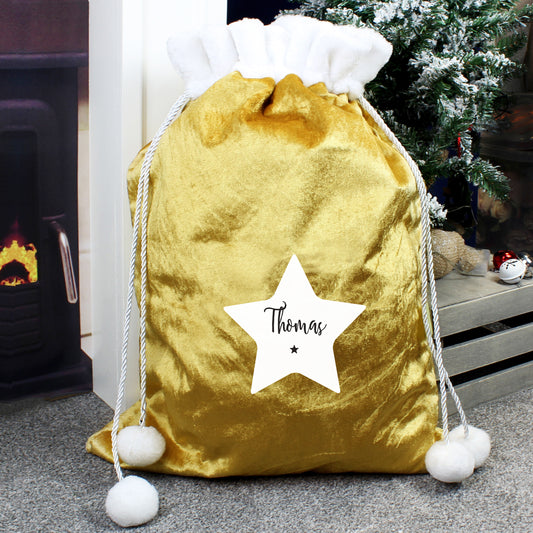 Personalised Gold velvet Christmas bag with a star and 'Thomas' text, placed in front of a fireplace and Christmas tree.
