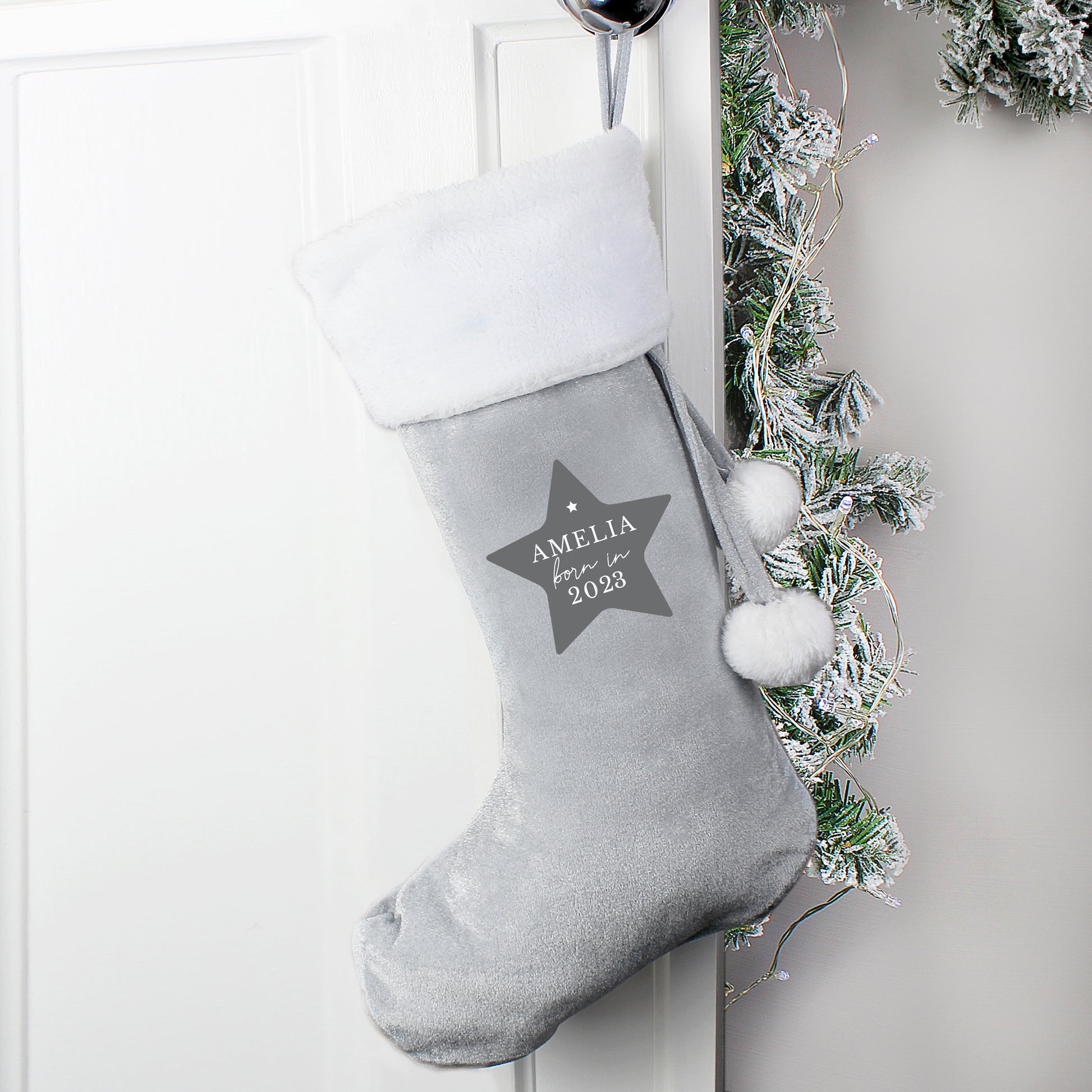 Gray Christmas stocking with a star design and text, hanging on a white door with greenery.