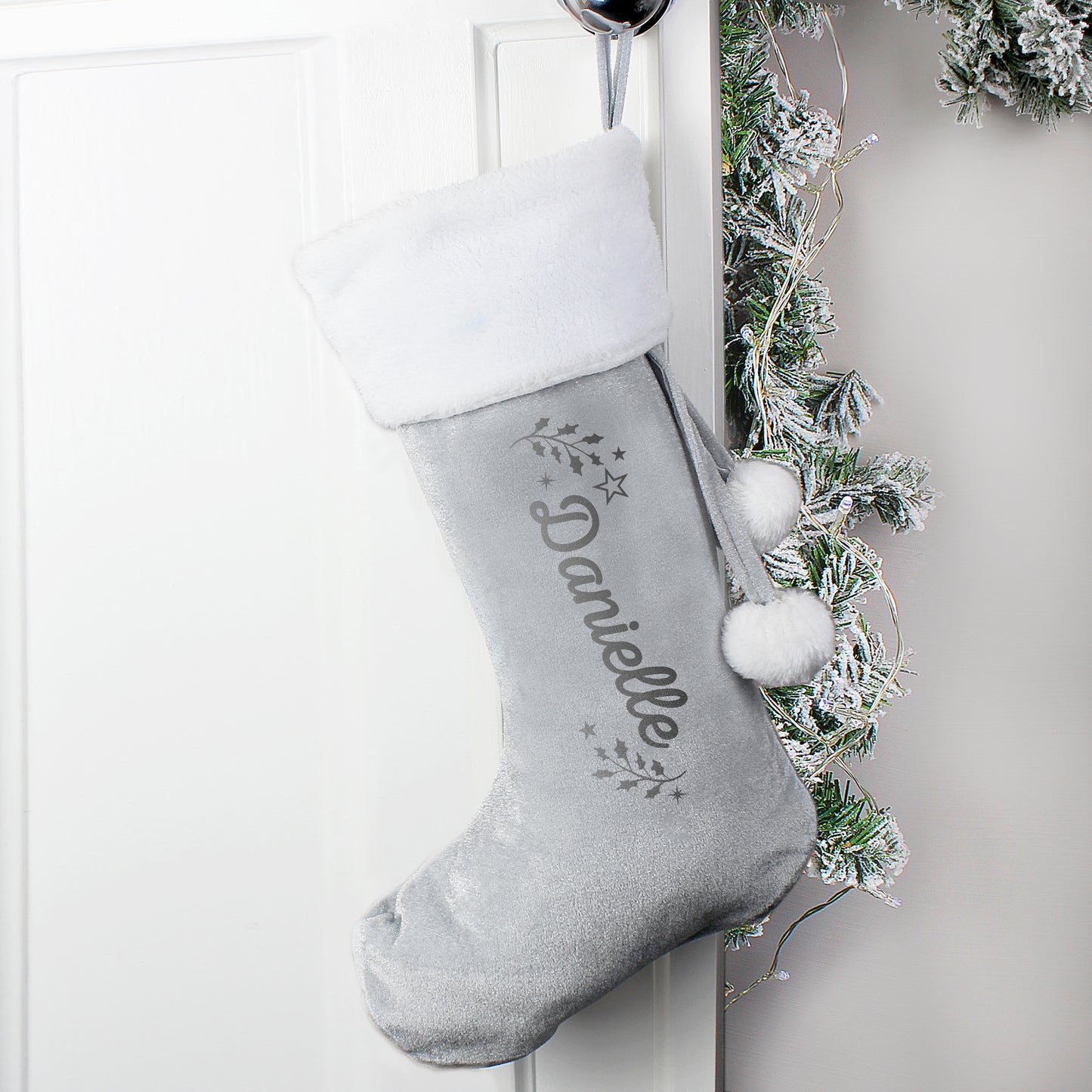 Grey Christmas stocking with white pom-poms and embroidered name 'Danielle' hanging on a white door with greenery.