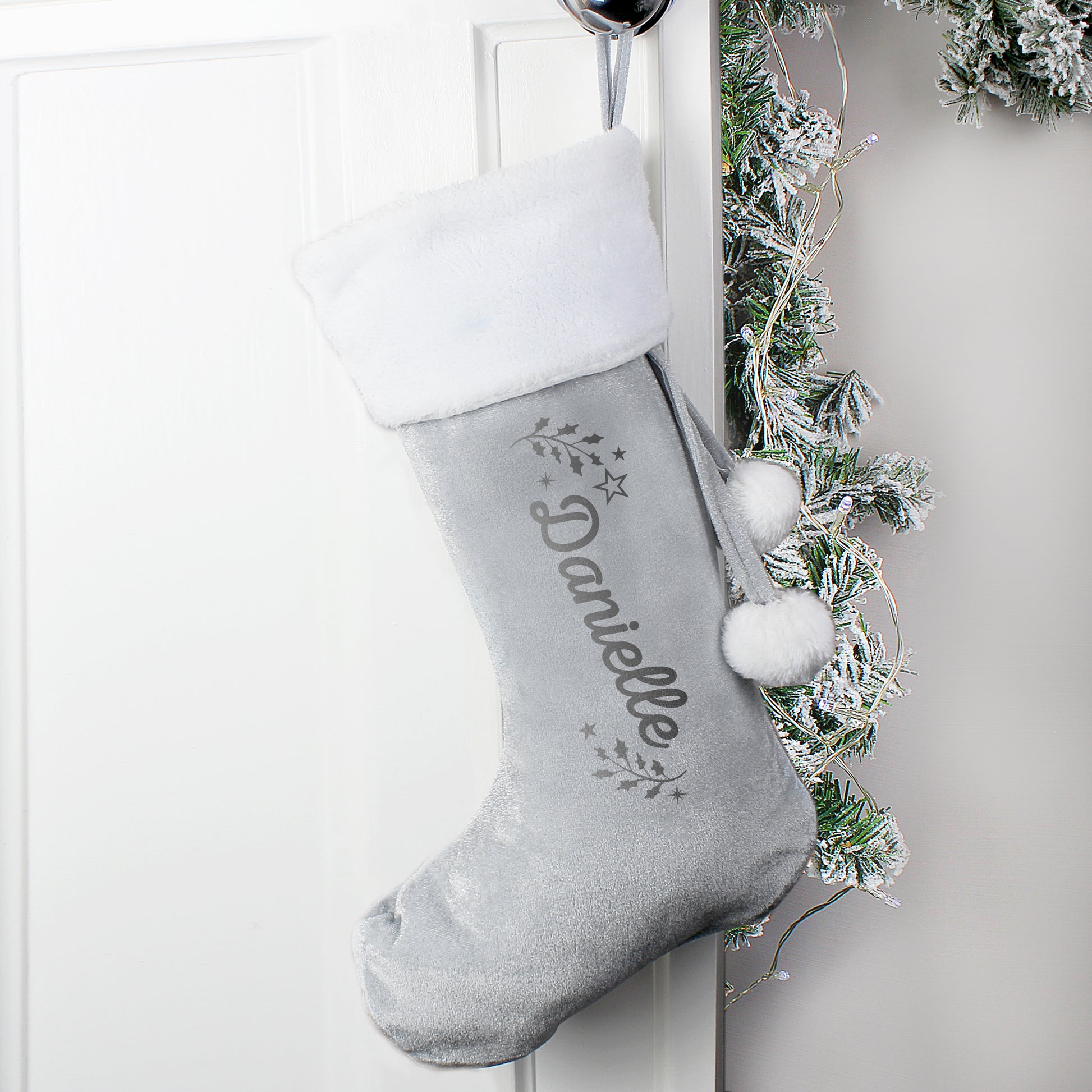 Grey Christmas stocking with white pom-poms and embroidered name 'Danielle' hanging on a white door with greenery.
