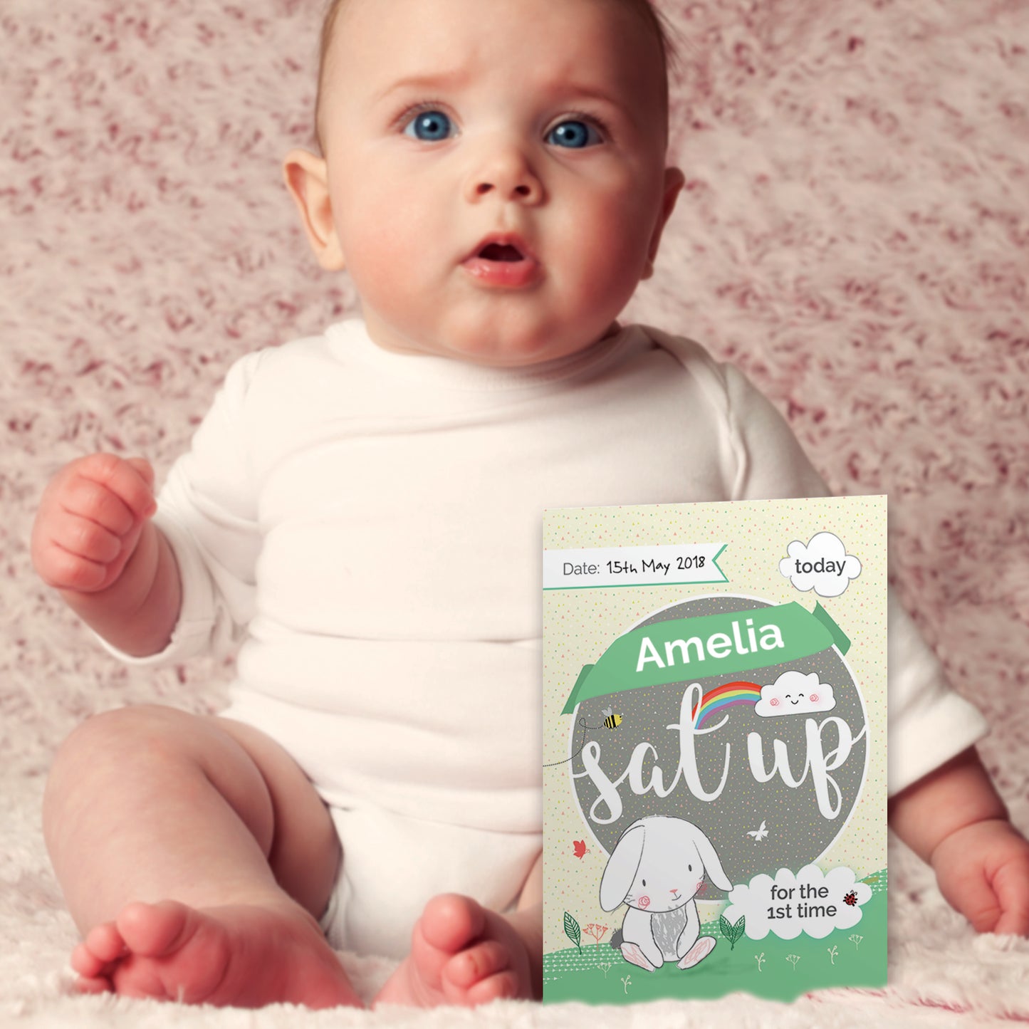 Baby sitting on a pink blanket with a milestone card indicating 'Amelia sat up for the 1st time'.