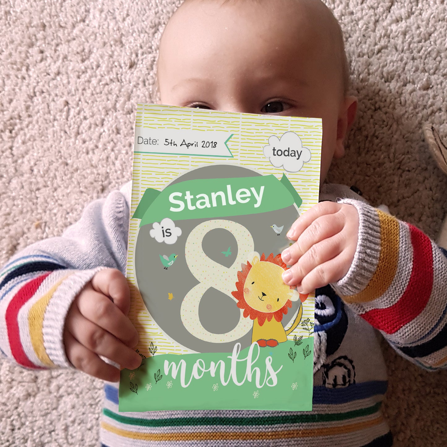 Baby holding a milestone card with 'Stanley is 8 months' on a carpeted floor.
